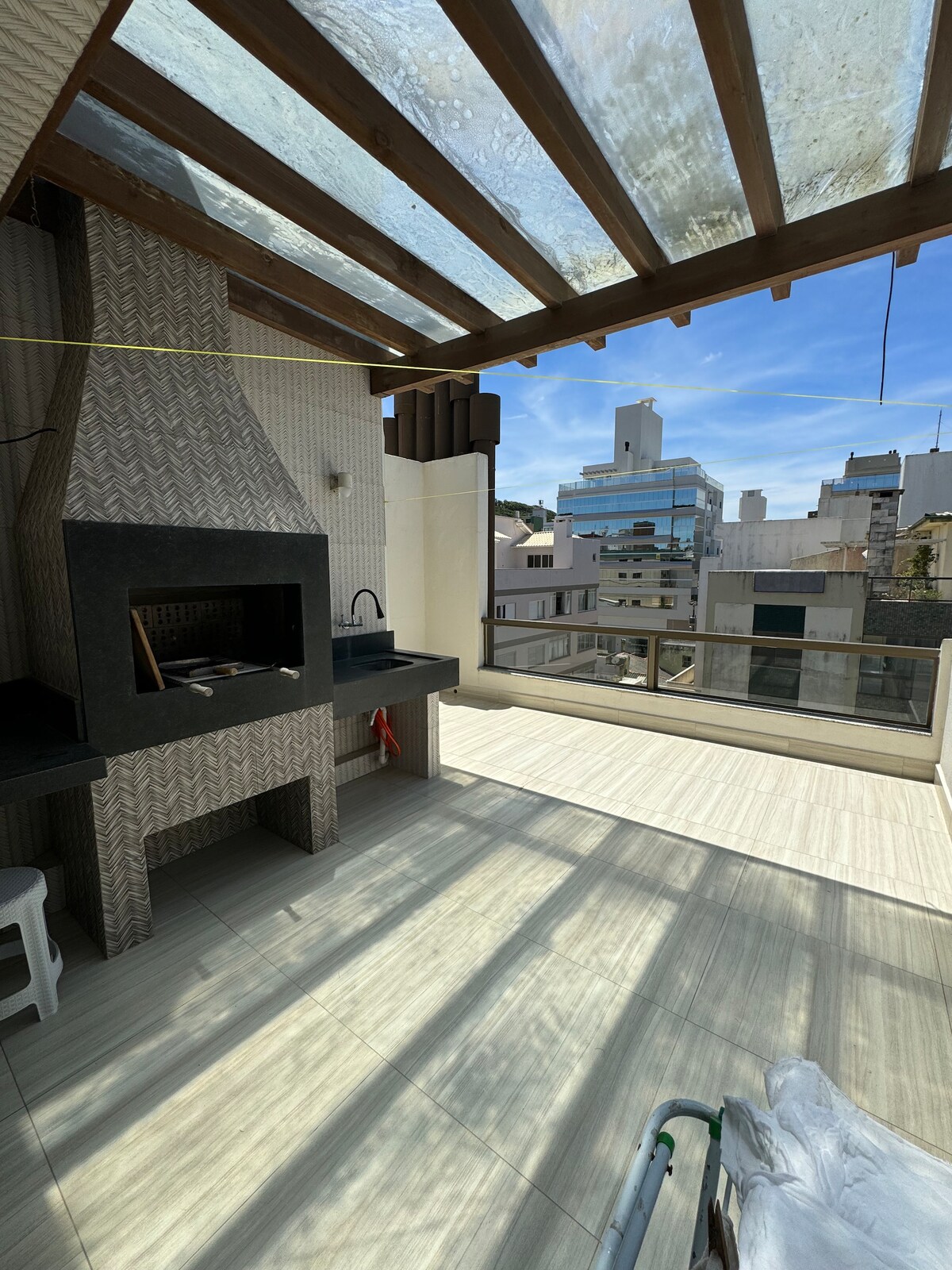 A spacious terrace features a built-in barbecue area, complemented by a sink and a stool. Sunlight filters through a clear roof, casting patterned shadows on the tiled flooring. The background reveals urban buildings, enhancing the outdoor experience.