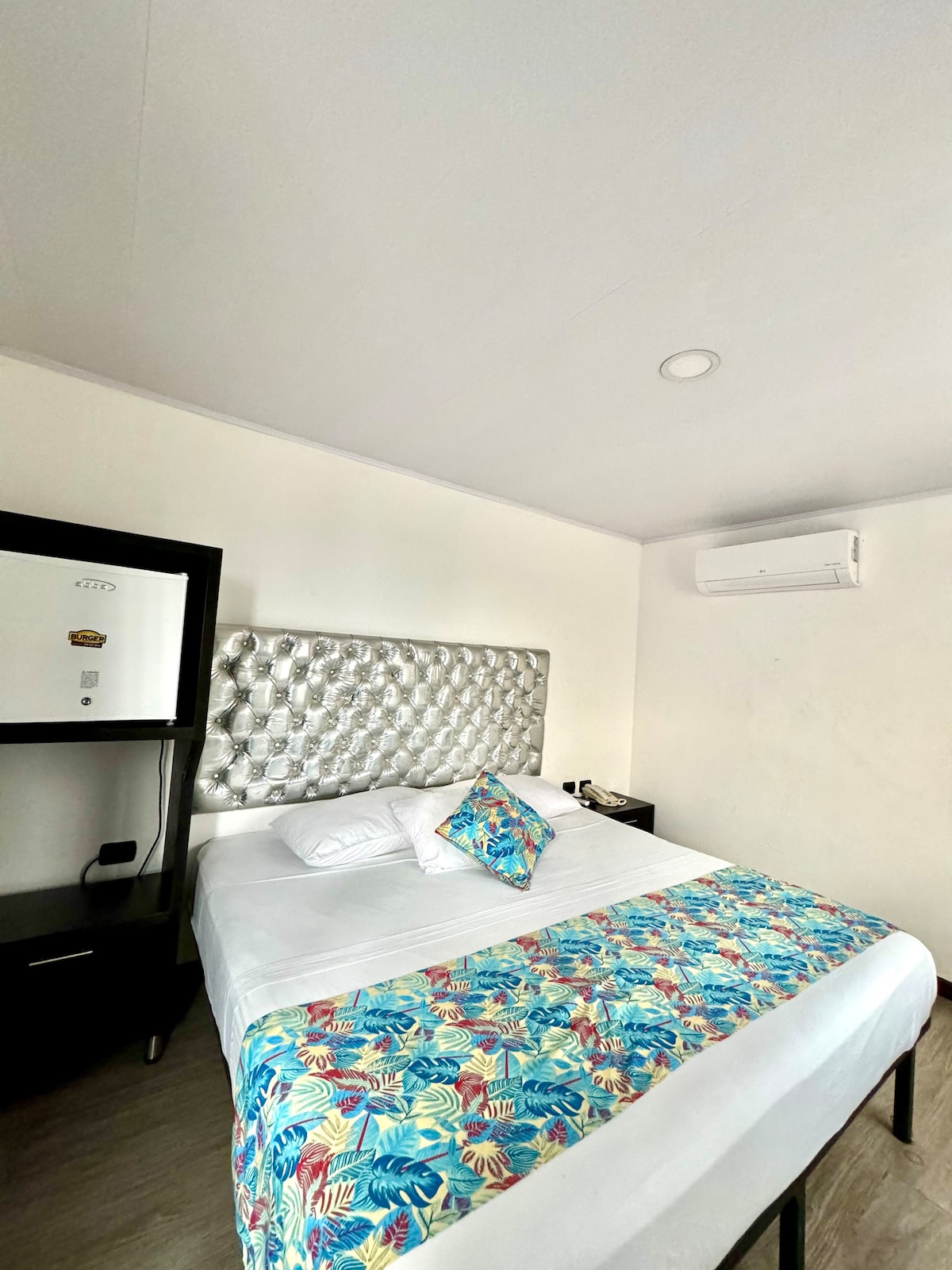A sleeping area features a bed with a textured headboard and a colorful tropical-patterned blanket. A small nightstand and a modern cabinet are visible, against minimalist white walls. An air conditioning unit is mounted on the wall for climate control.