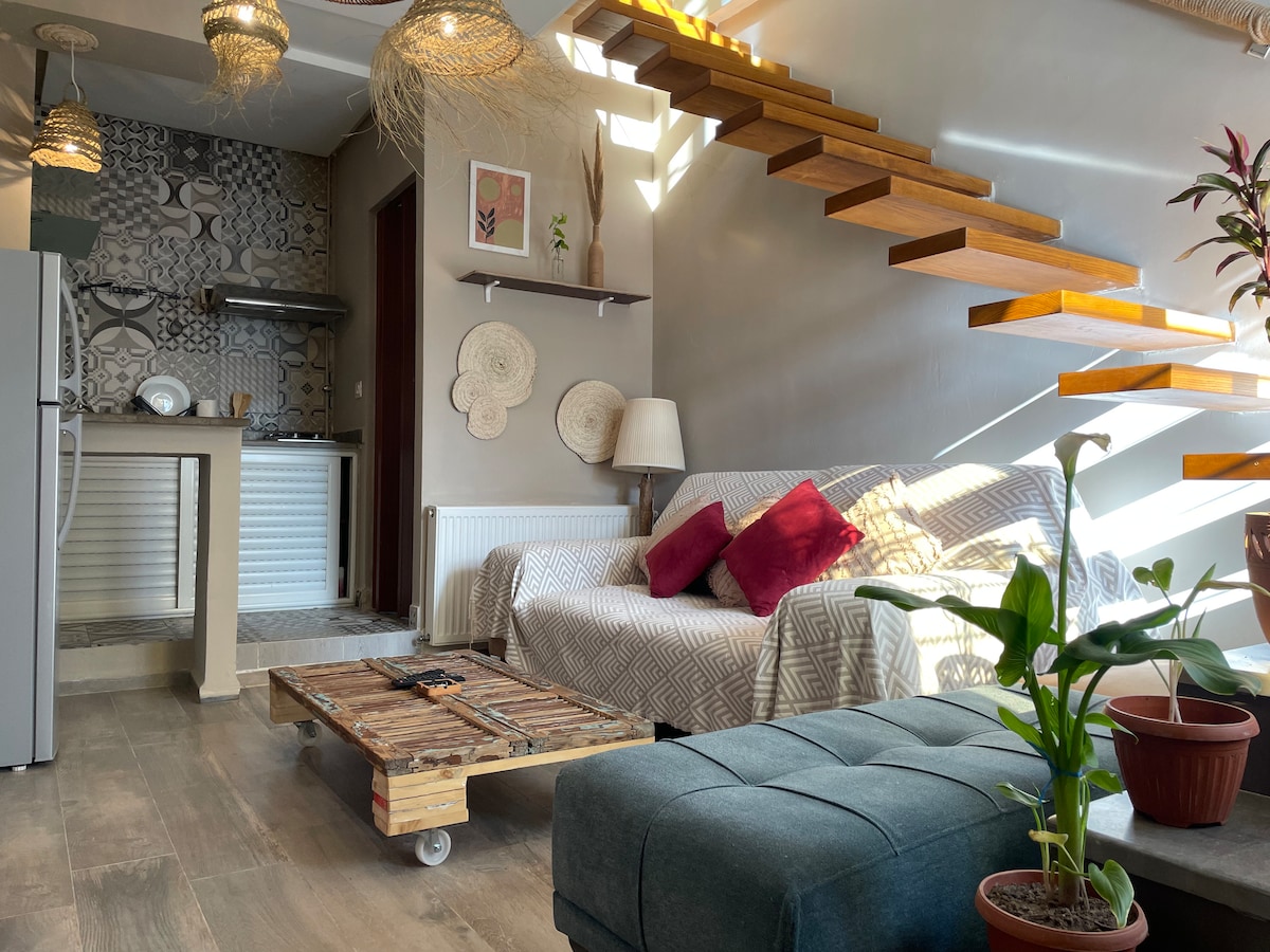 A welcoming living area is presented, featuring a light-colored couch adorned with decorative cushions. A wooden coffee table on wheels sits in front of the couch, while a potted plant adds a touch of greenery. A staircase with wooden steps leads to the upper level, illuminated by natural light.