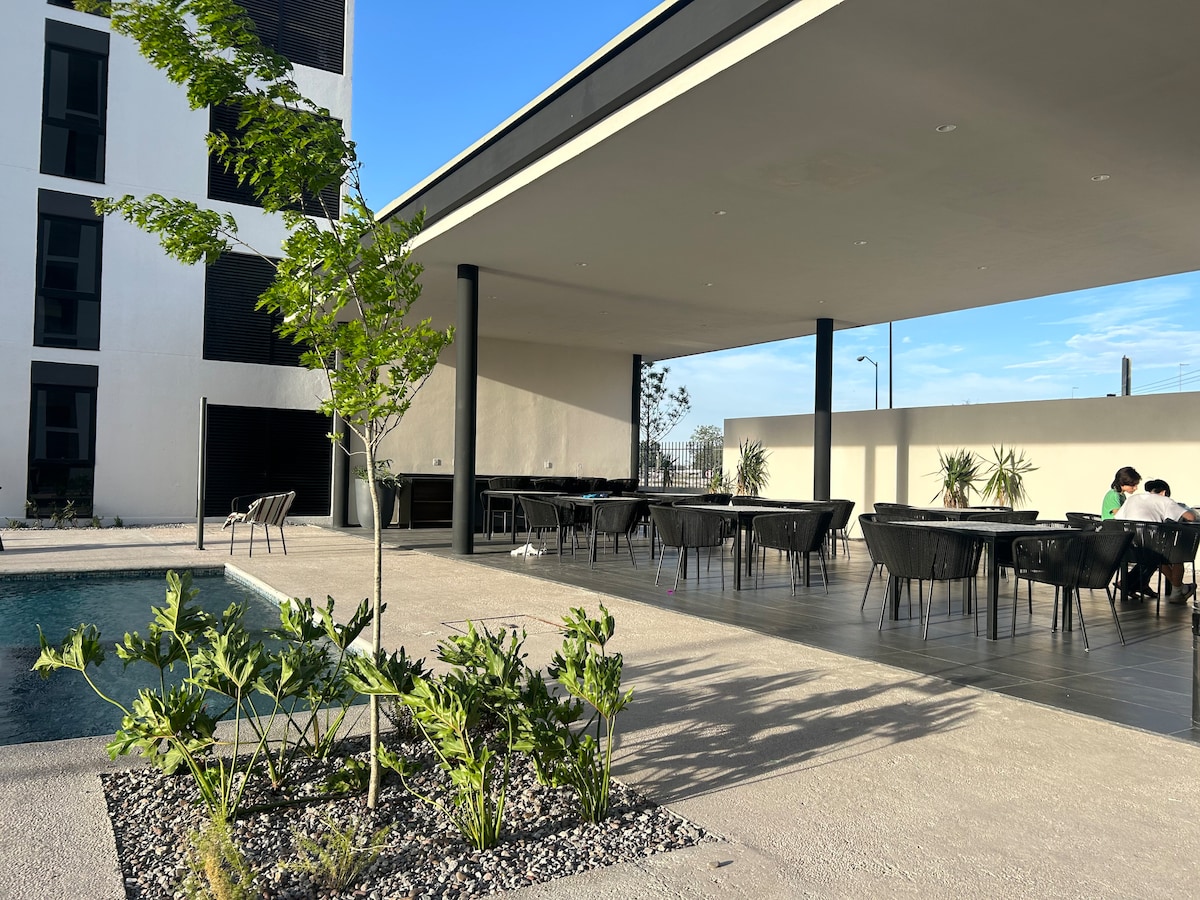 An outdoor area is depicted with a swimming pool and a shaded seating space. Modern chairs are arranged around tables, surrounded by greenery. The clean lines of the structure provide a sense of spaciousness, complemented by a bright blue sky.