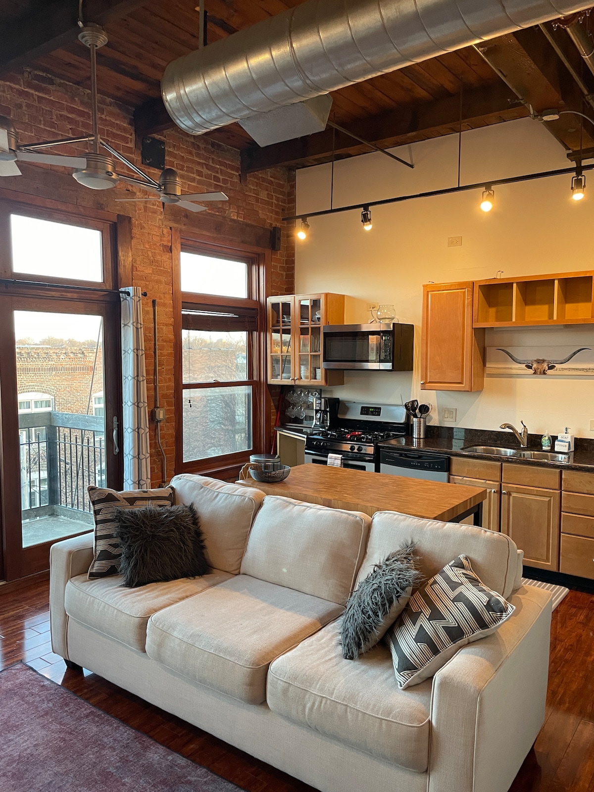 An open-plan living area is highlighted by exposed brick walls and wooden ceiling beams. A light-colored sofa with decorative cushions faces an area with a kitchenette and dining space. Large windows allow ample natural light, and outdoor views are visible from the upper level.