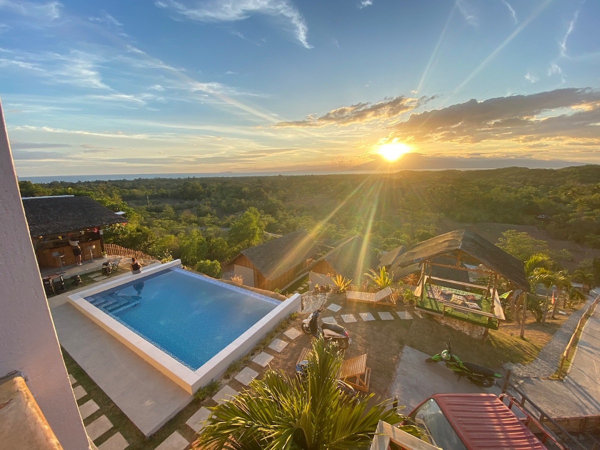 A spacious pool is set against a backdrop of lush greenery, with lounge chairs positioned nearby. The sun sets over the horizon, casting warm light across the landscape. Native cottages with thatched roofs are visible, surrounded by trees and open space.