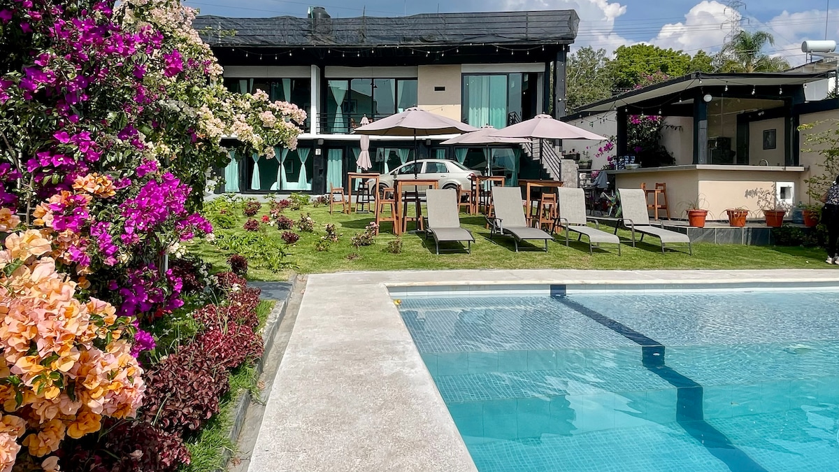 A lush garden features vibrant flowers surrounding a clear swimming pool. Sun loungers and shaded tables are arranged on the lawn, with two floors of the villa visible in the background, showcasing large balcony windows that invite natural light.