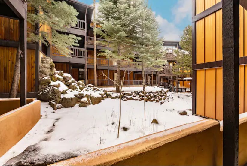 A view of the courtyard from a patio is presented, featuring snow-covered ground and several evergreen trees. Wooden balconies and stone features are visible in the background, adding to the natural setting of the condo complex.