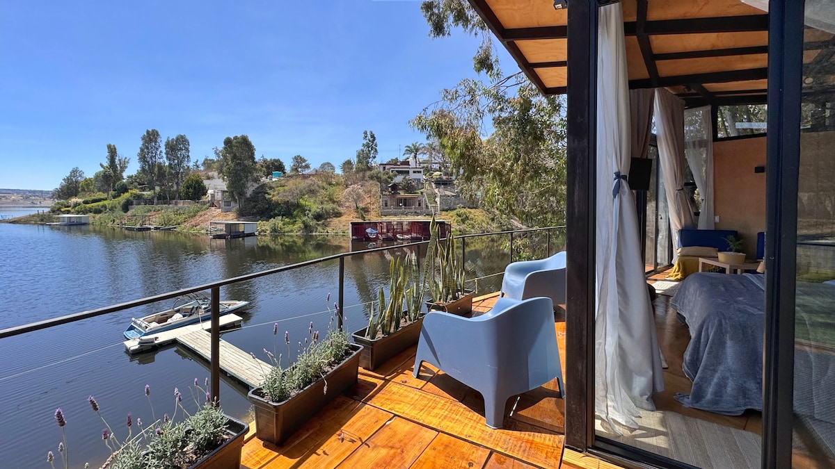 An outdoor terrace offers a view of the lake, featuring two blue chairs positioned beside potted plants. The cabin's interior is visible, showcasing a blue bedspread and a flat-screen TV mounted on the wall. The calm water reflects the surrounding scenery.