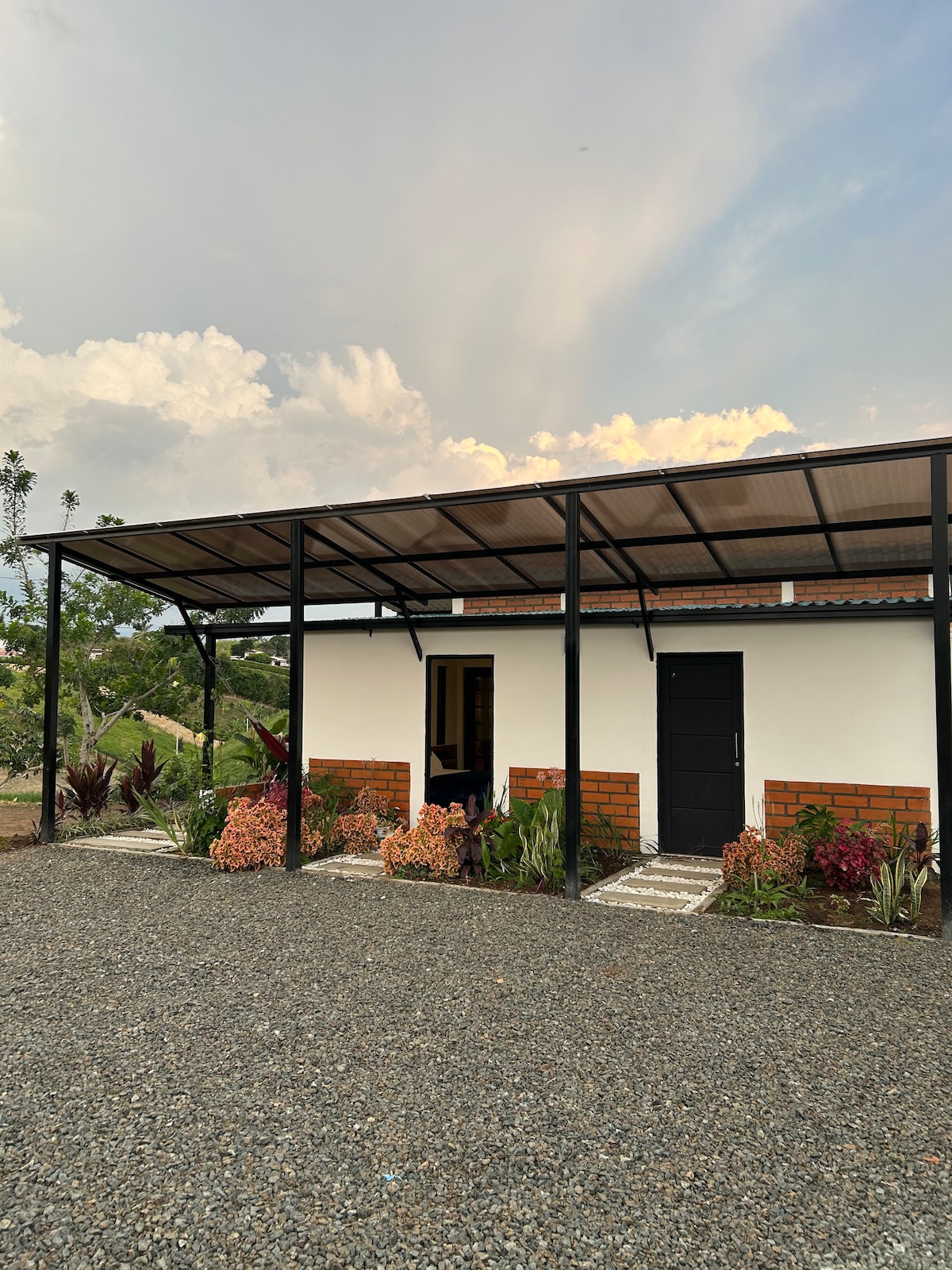A modern cabin features a covered patio with landscaping of colorful plants and gravel paths. The exterior walls are painted white, with contrasting brick accents. A single door leads into the cabin, while a large overhang provides shade, creating a comfortable entrance.