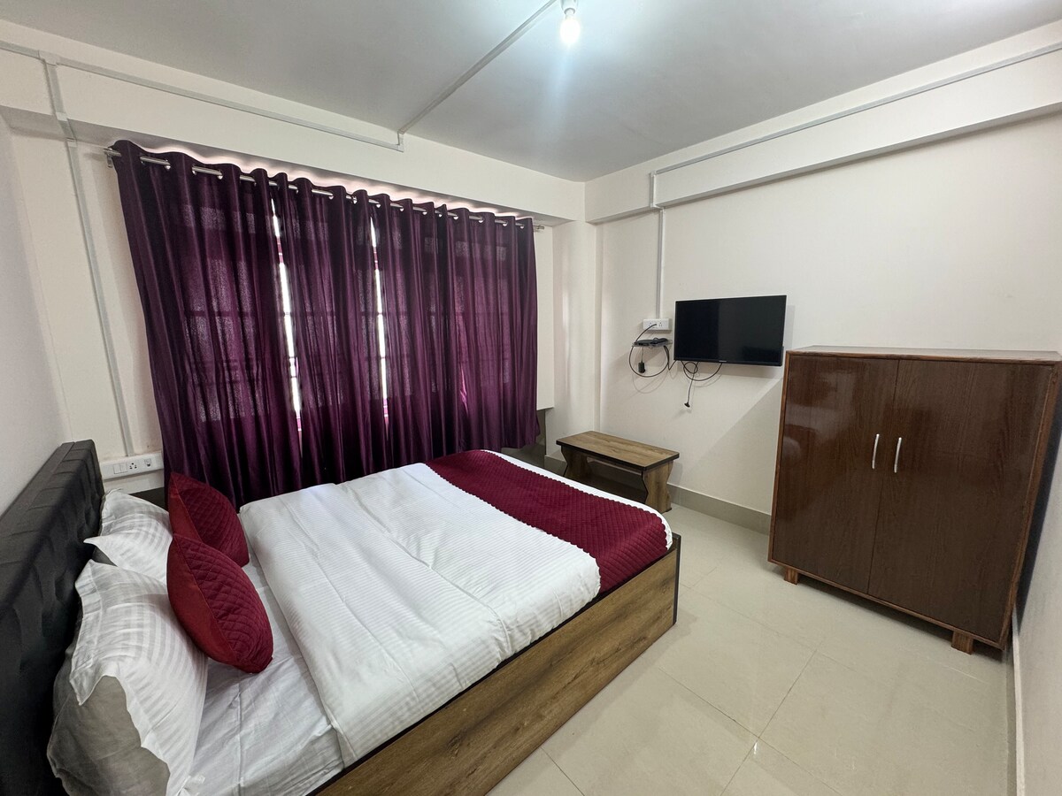 A bedroom is presented with a spacious bed dressed in crisp white linens, accented by deep red pillows. Dark purple curtains frame the window, while a wooden dresser and a small table complement the minimalist decor. A wall-mounted television offers entertainment options.