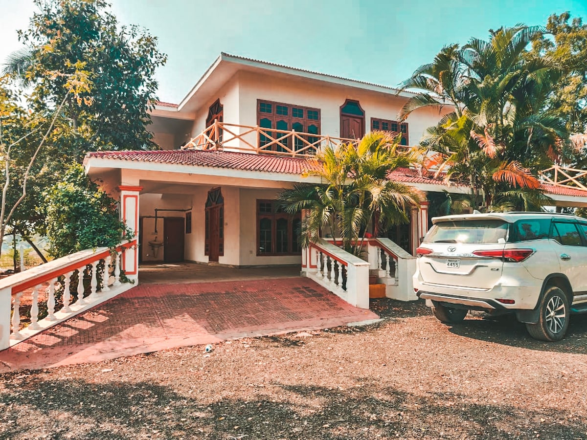 A spacious two-story house is surrounded by lush greenery, showcasing a welcoming front porch with decorative railings. A white vehicle is parked in the driveway, with vibrant plants framing the entrance. Large windows allow for natural light to brighten the exterior.