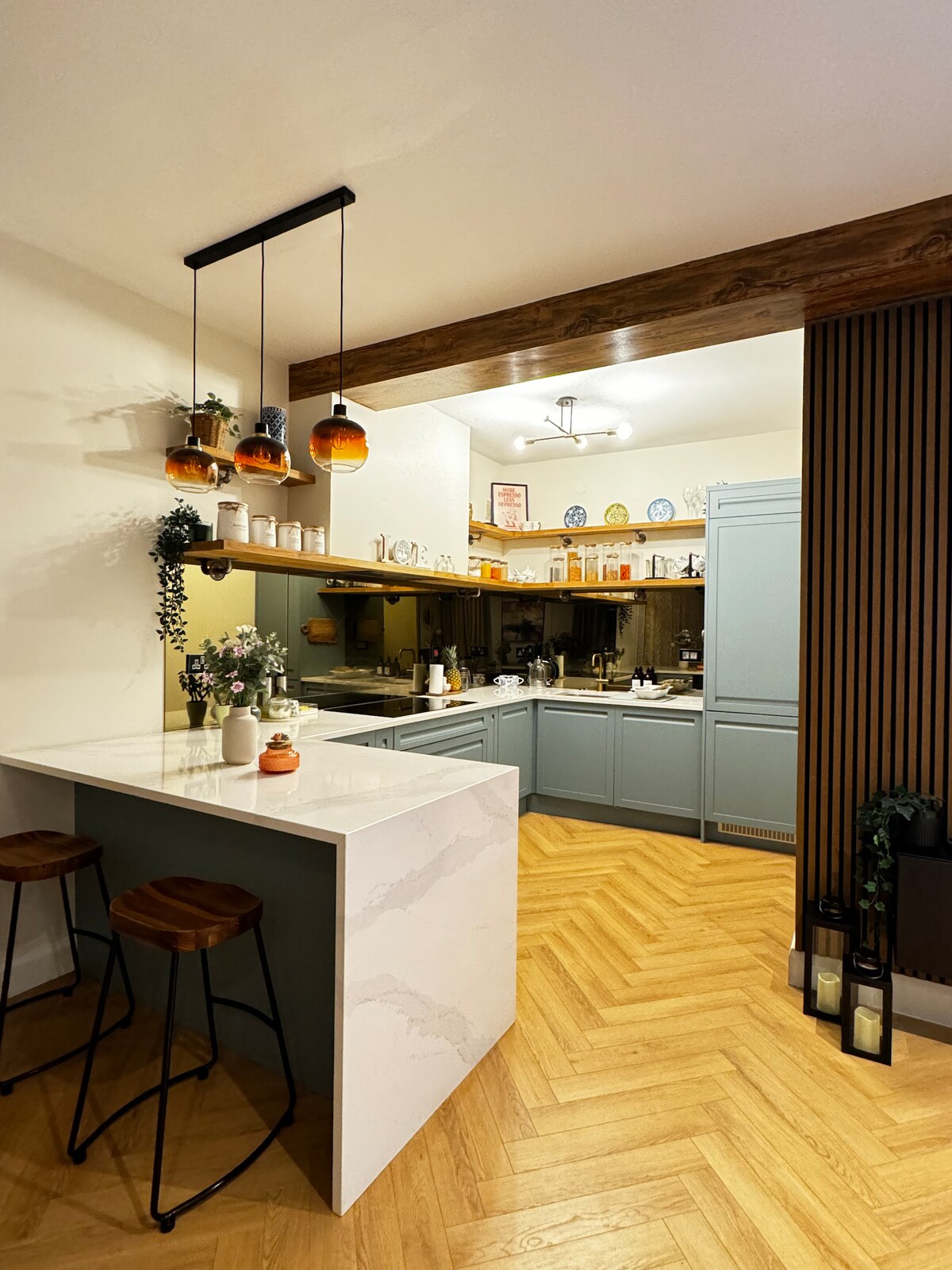 A stylish kitchen space features a marble-topped island with two wooden stools. Custom cabinetry in soft blue complements the chevron-patterned wooden flooring, while pendant lights with orange shades add a warm touch. Decorative plates line the shelves, providing subtle color against the backdrop.