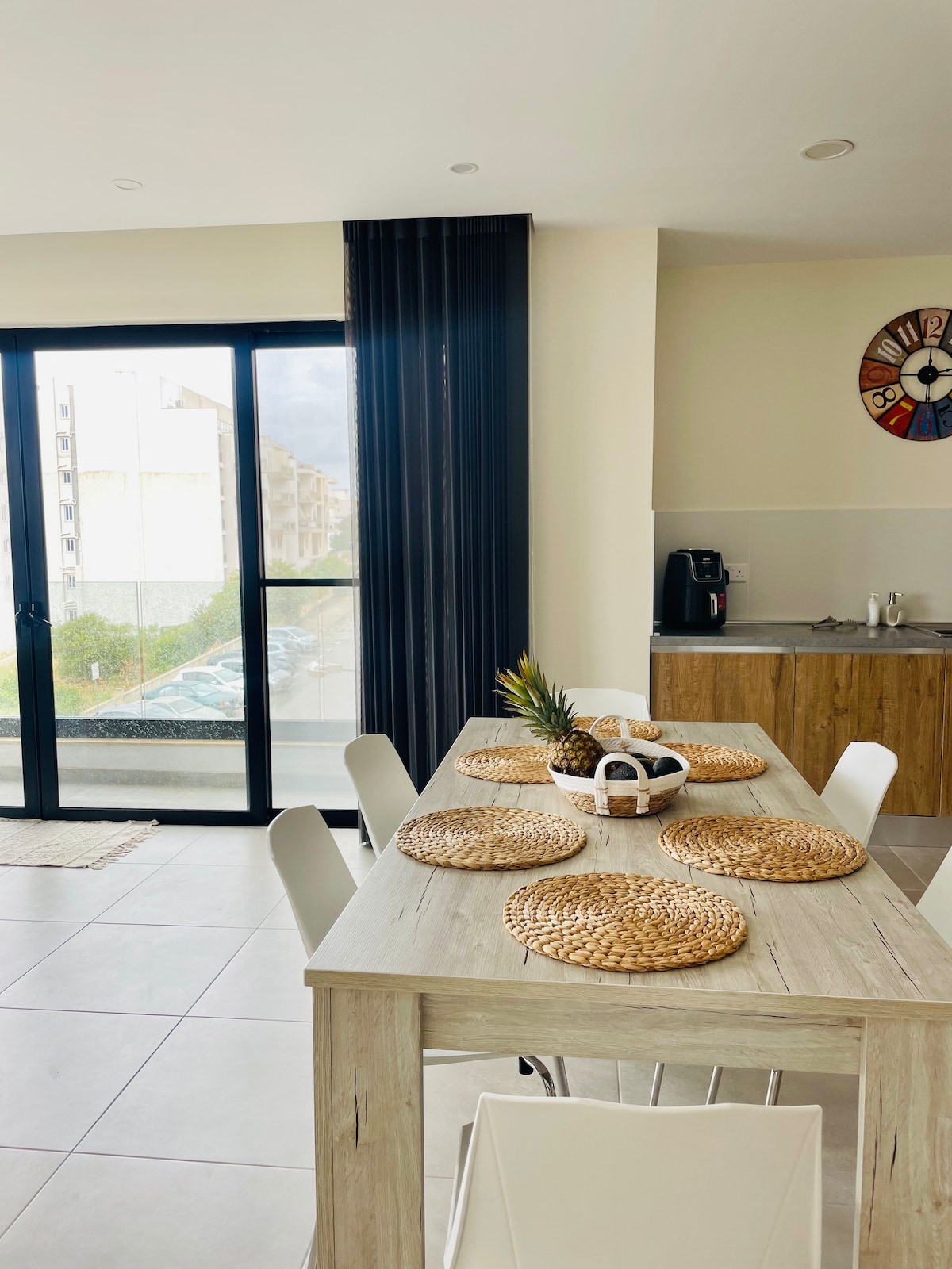 A modern dining area is presented with a spacious wooden table, adorned with round woven placemats and a central decorative arrangement. Two large glass doors provide views of the outdoors, while a compact kitchen area features wooden cabinetry and a coffee station.