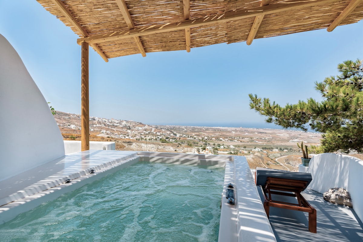 A private hot tub is set under a wooden pergola, surrounded by a spacious deck. Expansive views of the island's landscape and coastline are visible in the background, enhancing the feeling of seclusion. A wooden lounge chair is positioned beside the hot tub.