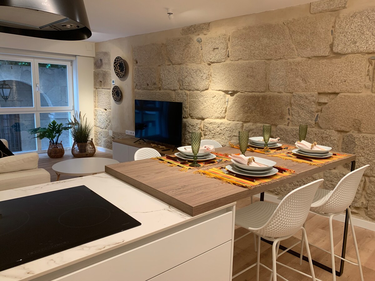A modern kitchen space features a central island with seating for four. Textured stone walls provide a warm backdrop, complemented by a large screen TV and decorative elements. Natural light filters in through a large window, enhancing the inviting setup of tableware on the island.