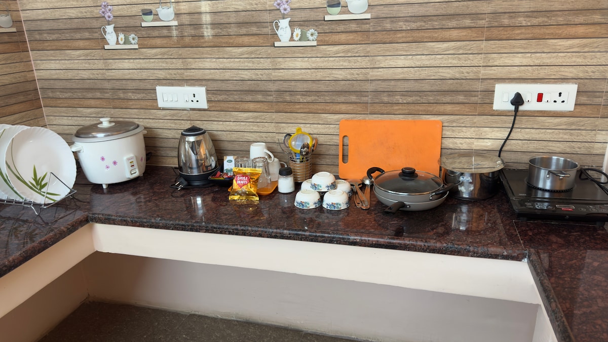 A functional kitchen space is showcased, featuring essential utensils and appliances. A rice cooker, kettle, and frying pan are present, alongside cutting boards and various dishes. The countertop is streamlined with a subtle color palette, offering ample room for food preparation.