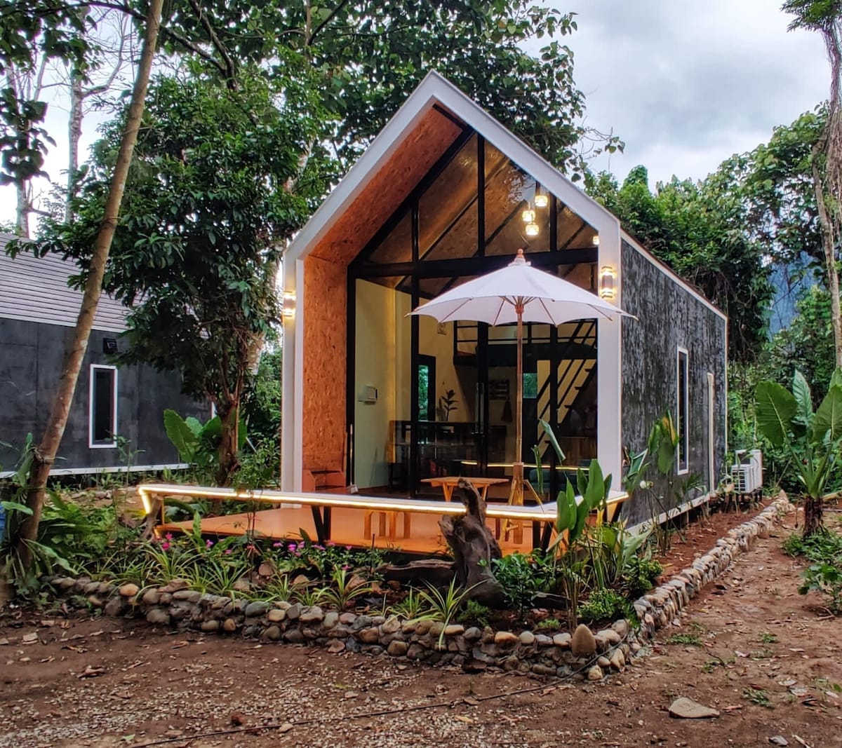 A modern cabin features a distinctive gabled roof with large glass windows. The exterior is framed by lush greenery and decorative stones. A small patio area is equipped with an umbrella and surrounded by tropical plants, inviting guests to enjoy the serene outdoor space.