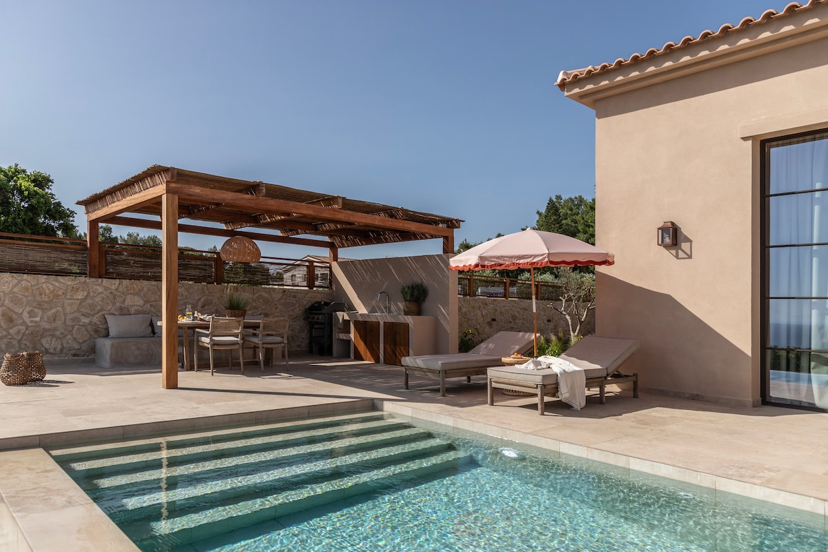An outdoor area features a private infinity pool with clear water, surrounded by sun loungers. A shaded dining space with a wooden pergola offers an inviting setting. A barbecue area is visible, complemented by subtle greenery and rustic stone walls.