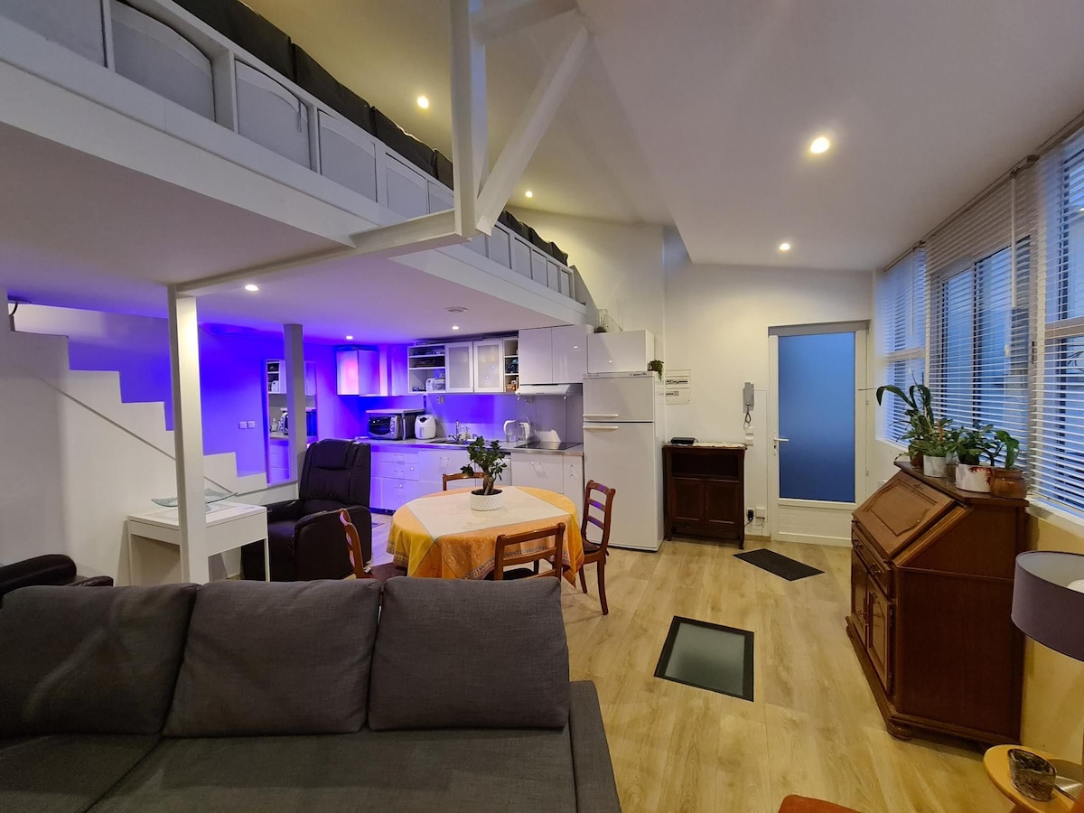 A spacious loft layout is visible, featuring a kitchen with modern appliances and a dining area with a round table. Soft lighting highlights the space, while a staircase leads to the mezzanine. Large windows allow natural light, and wooden accents are present throughout.