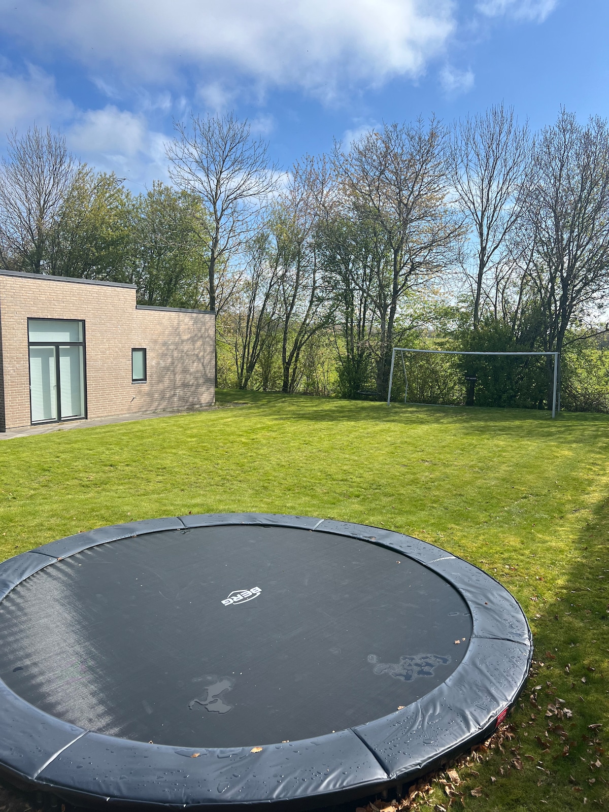 An outdoor space features a large trampoline centered on a neatly manicured lawn. In the background, a goalpost stands ready for play, framed by a border of trees under a partly cloudy sky. The area is spacious, inviting outdoor activities.
