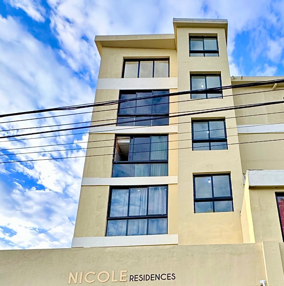 The exterior of the building is depicted with a modern design, featuring a muted beige facade and large windows that invite natural light. The name 'Nicole Residences' is displayed prominently at the base, while a clear sky and scattered clouds serve as the backdrop.