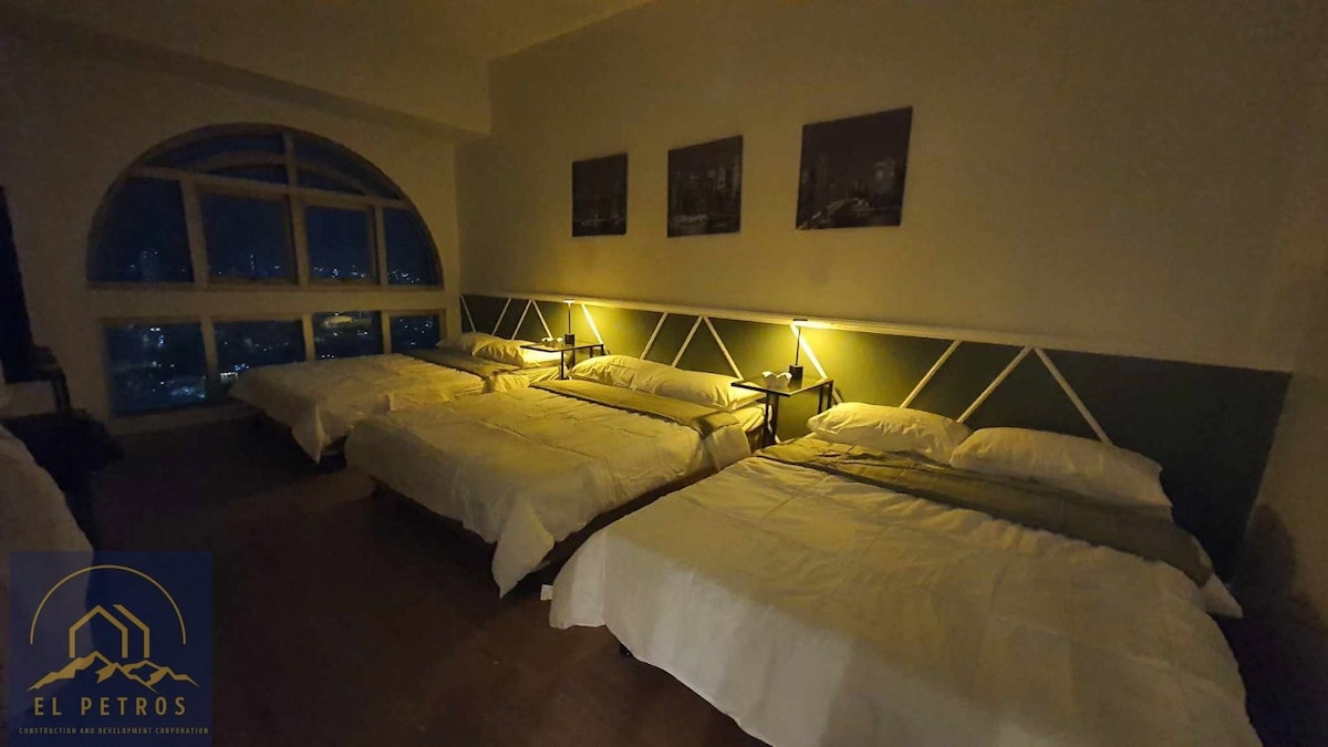 A spacious sleeping area is presented with three double size beds, each neatly arranged with white linens. Soft lighting from bedside lamps casts a warm glow, enhancing the calm atmosphere. Large windows offer views of the city skyline, and minimalist wall art adds subtle character.