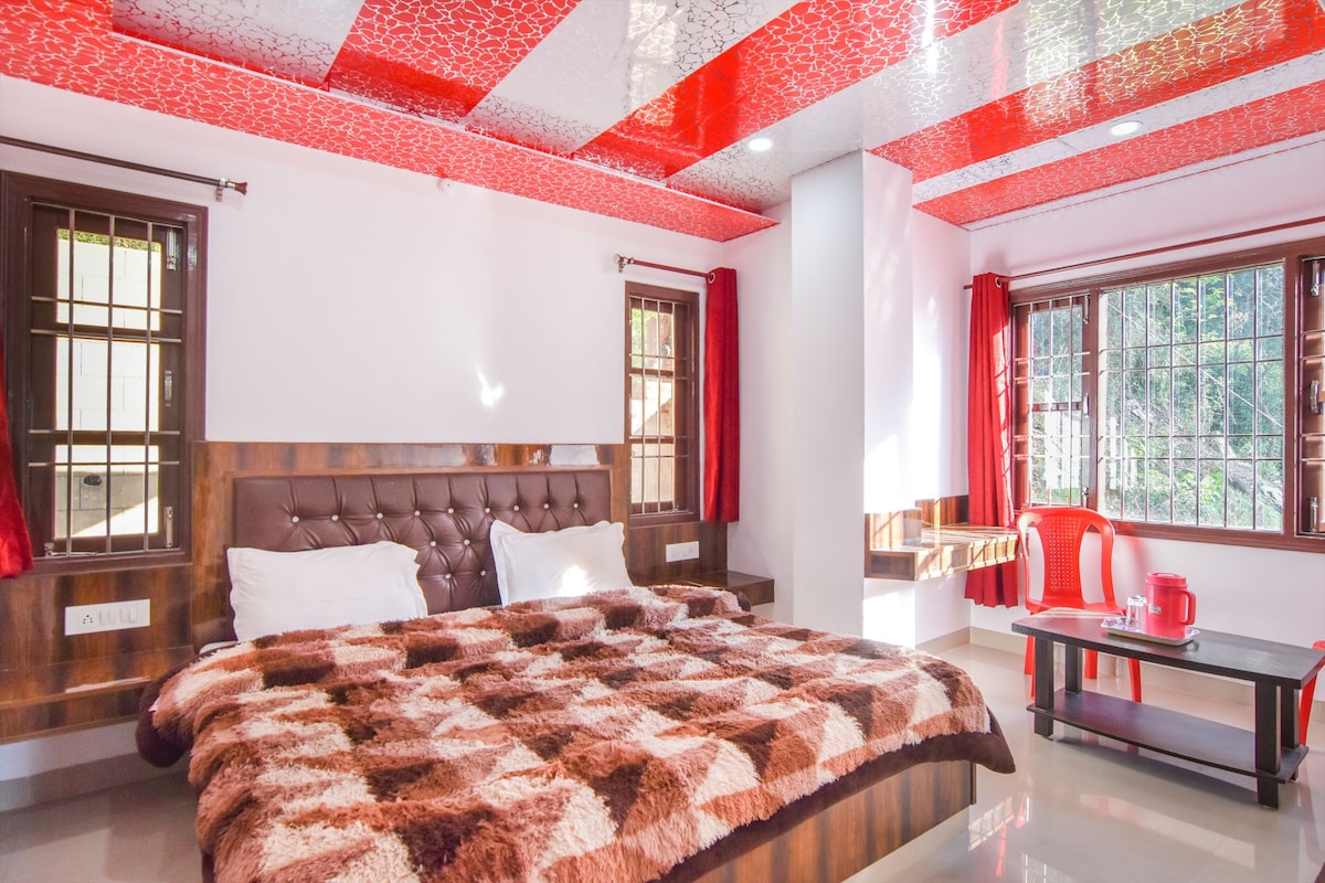 A well-lit room features a spacious bed with a patterned blanket, accompanied by two red chairs and a small table. Large windows with grids allow natural light to fill the space, while bright red curtains complement the ceiling design.