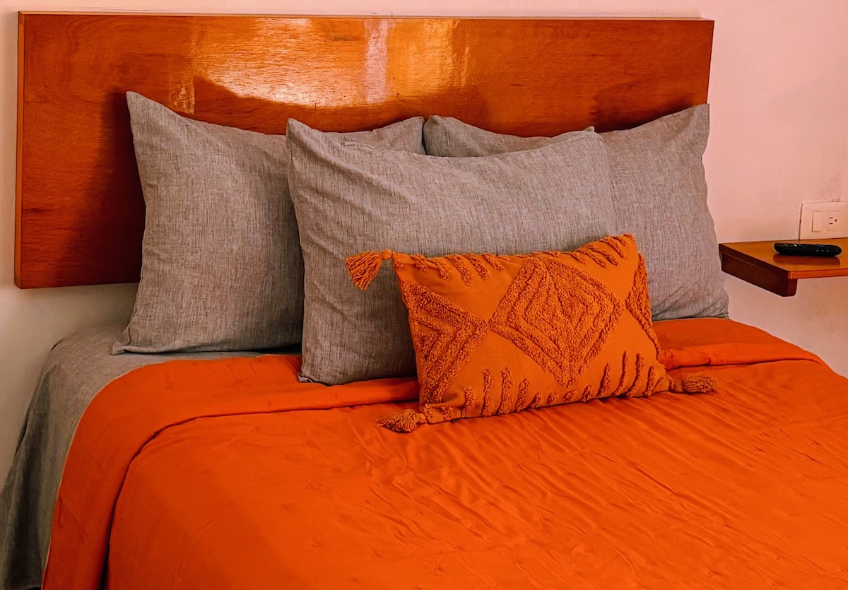 A spacious bed is adorned with a vibrant orange coverlet and accented by several gray and patterned decorative pillows. A wooden headboard offers a warm contrast, while a small bedside table holds a remote control.