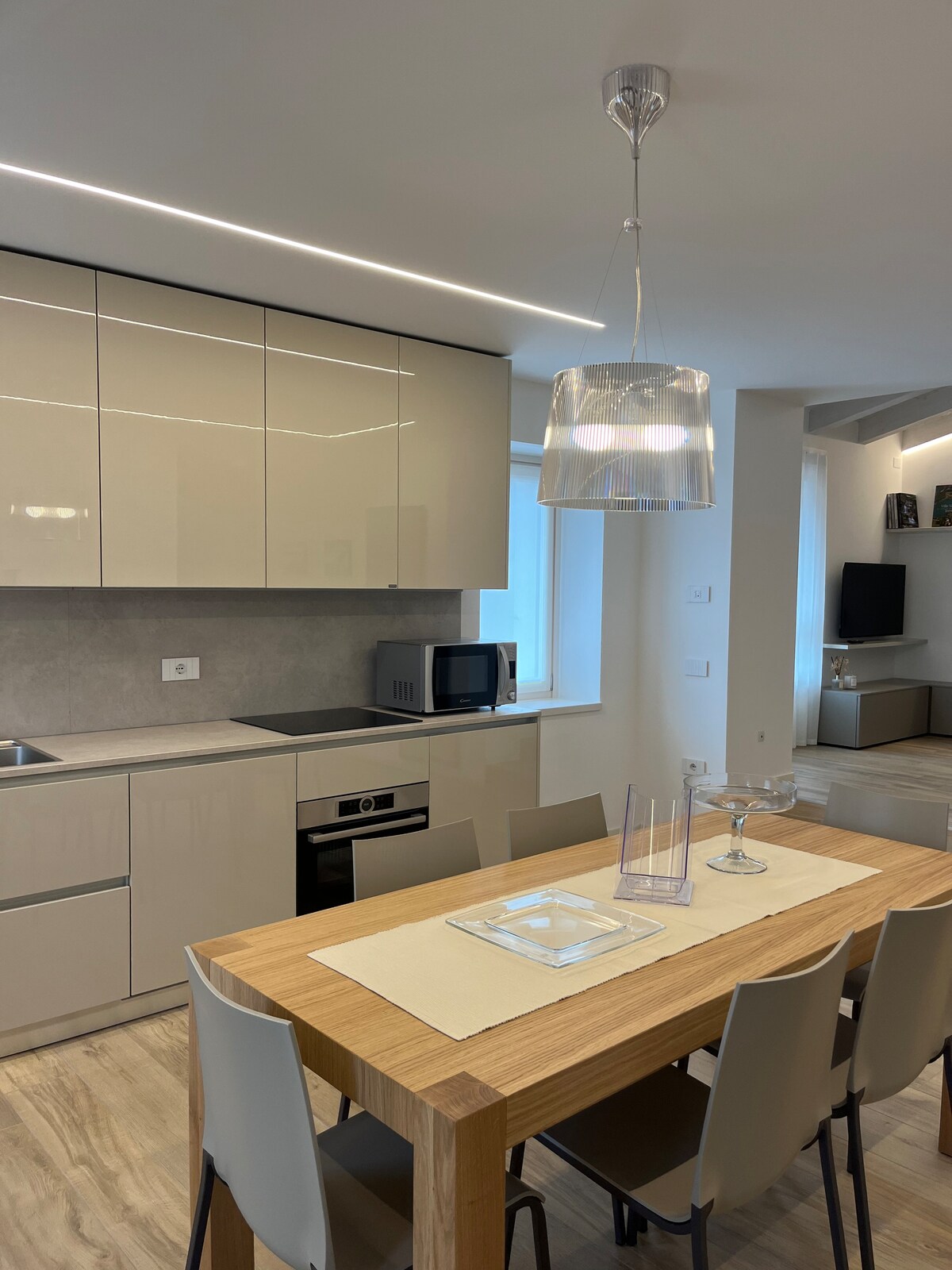 A modern kitchen and dining area hosts a wooden table surrounded by light-colored chairs. Glossy cabinets reflect natural light, while a pendant light hangs above the table. A microwave and other essentials are neatly arranged in the kitchen space, enhancing functionality.