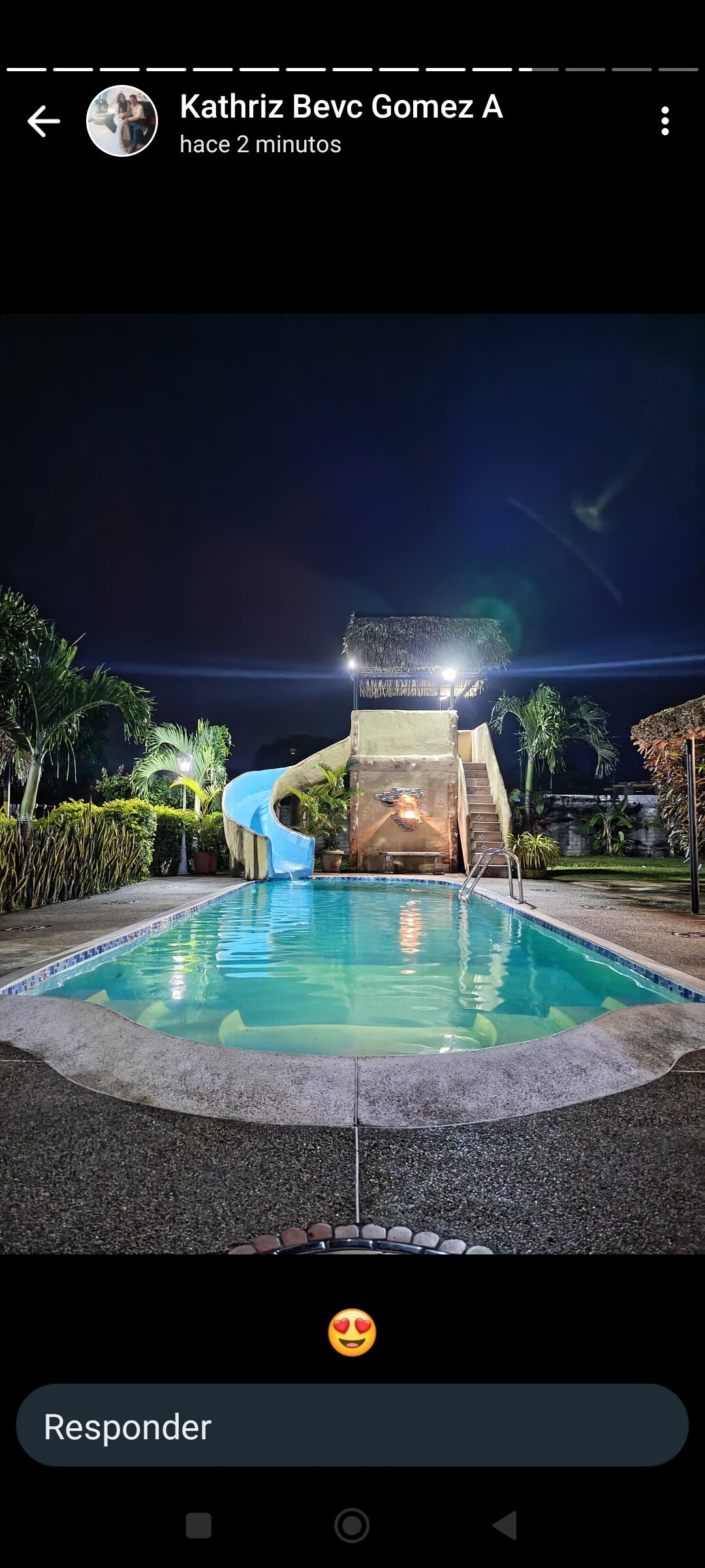 A sparkling pool area features a tall water slide on a stone staircase, surrounded by lush greenery. Soft lighting enhances the space, creating an inviting ambiance as evening approaches. The clear water reflects the surrounding landscape and the night sky.