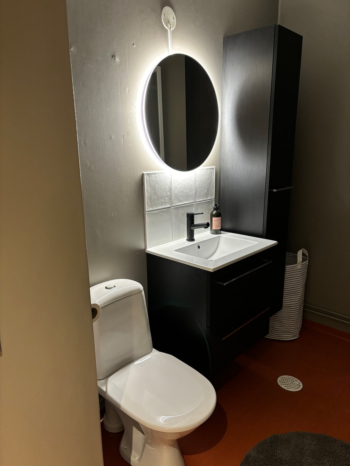 A compact bathroom features a modern toilet and a sleek vanity with a round, backlit mirror. A black faucet complements the white sink, and storage is provided by a dark cabinet above and a woven laundry basket nearby.