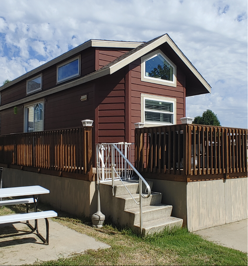 A tiny home is featured, characterized by a charming exterior with horizontal wooden siding and multiple windows. A wrap-around deck is visible, bordered by a wooden railing. Steps lead to the entrance, offering access to the inviting outdoor space.