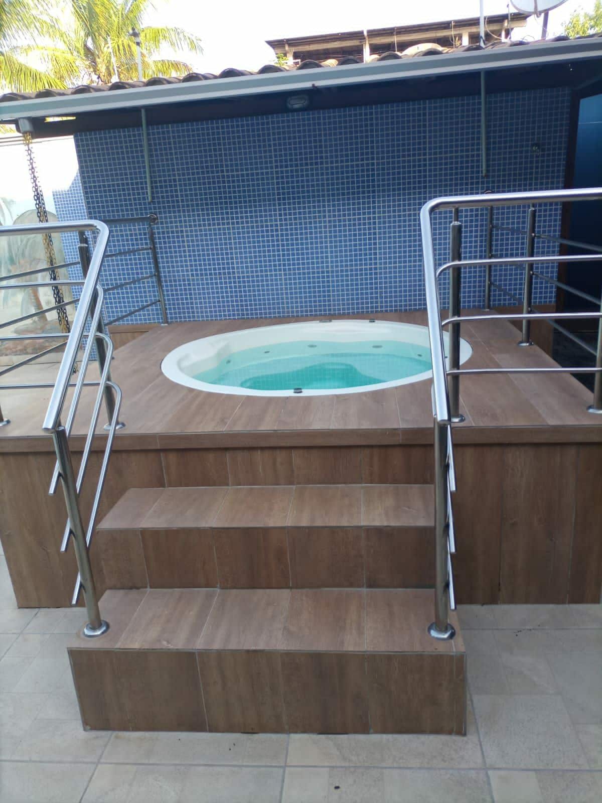 A raised jacuzzi is positioned on a wooden platform, surrounded by safety railings. Steps lead up to the jacuzzi, which features a smooth, round shape and clear blue water. Blue tiles adorn the wall behind, adding a clean and refreshing aesthetic.