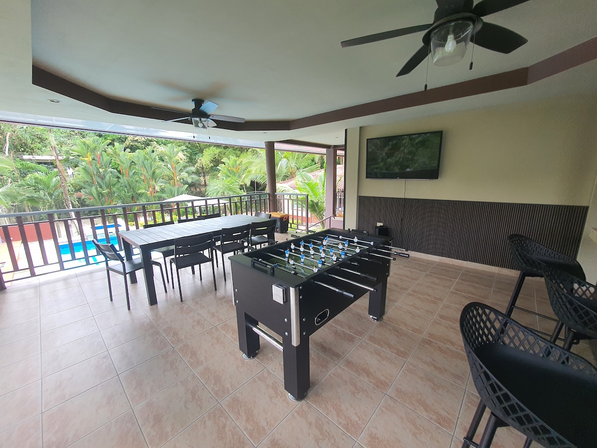 A spacious outdoor area is featured, equipped with a foosball table and a large dining table. Comfortable seating is arranged around the space, with a wall-mounted flat-screen TV visible. Lush greenery and a swimming pool are seen through the railing.