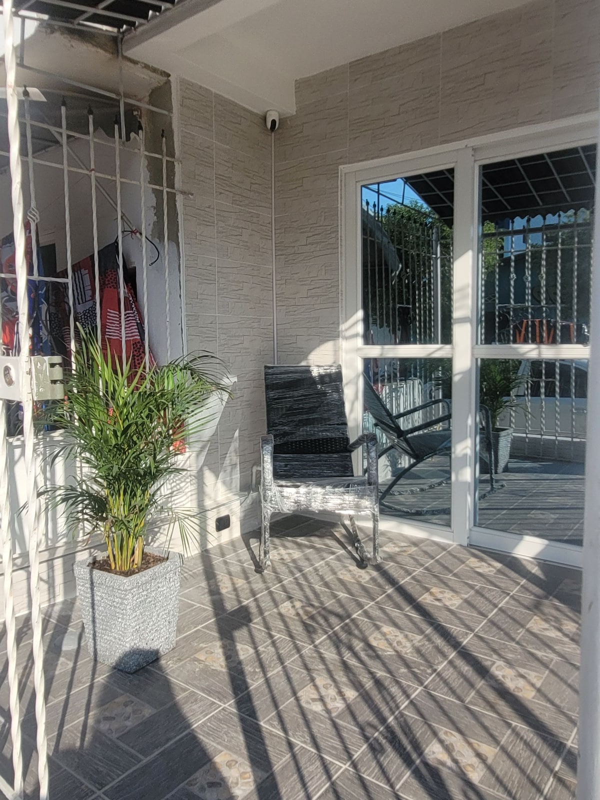 An inviting outdoor space features a potted plant next to a wicker chair, situated on a tiled surface. Sunlight casts shadows, creating a serene ambiance. The entrance is framed by large glass doors, providing a glimpse into the interior area.