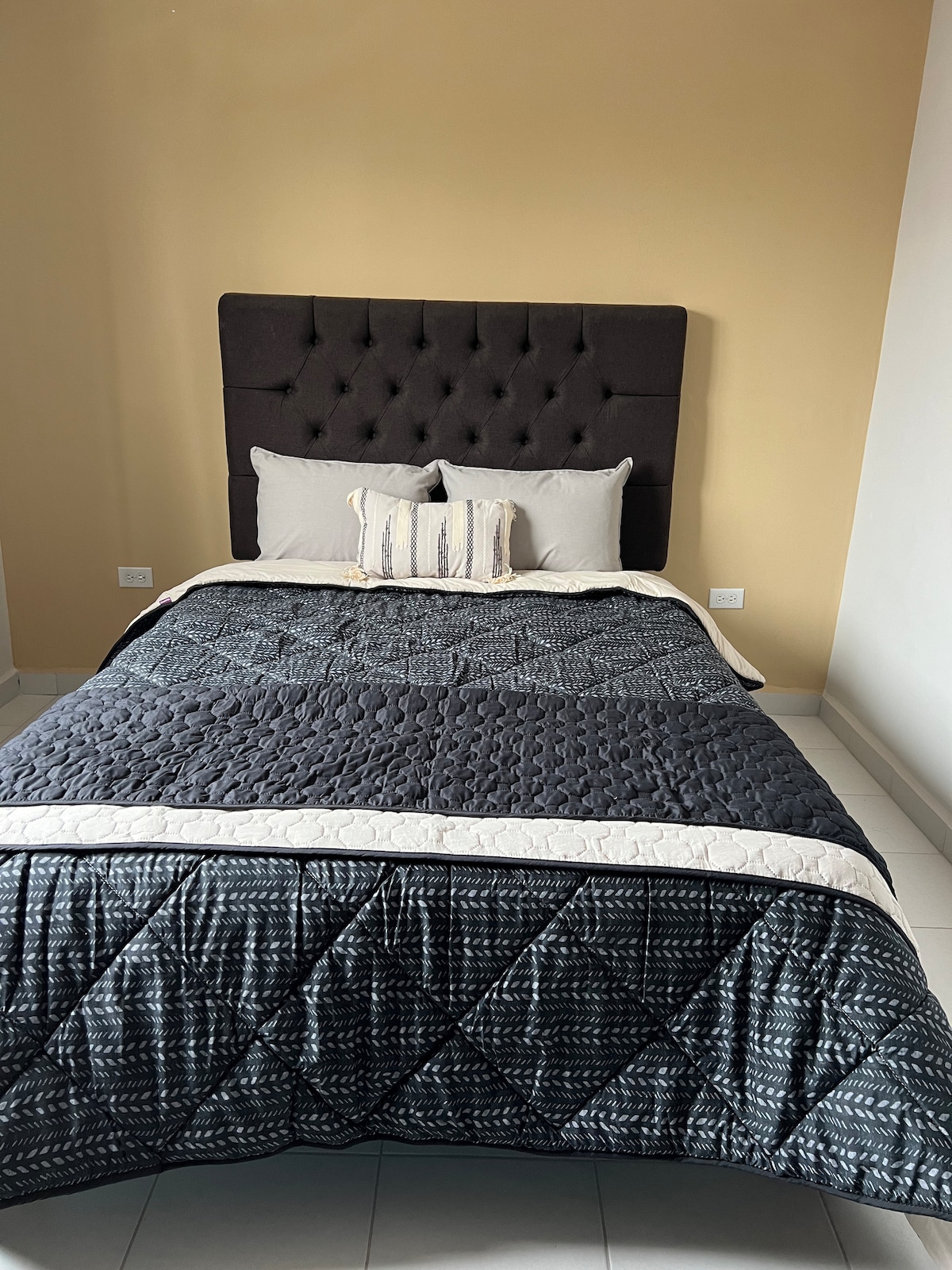 A cozy sleeping area features a dark upholstered headboard behind a neatly made bed with a patterned quilt. Soft, neutral-colored pillows complement the bedding. Light-colored walls create a calm backdrop, while floor tiles enhance the room's modern and clean aesthetic.