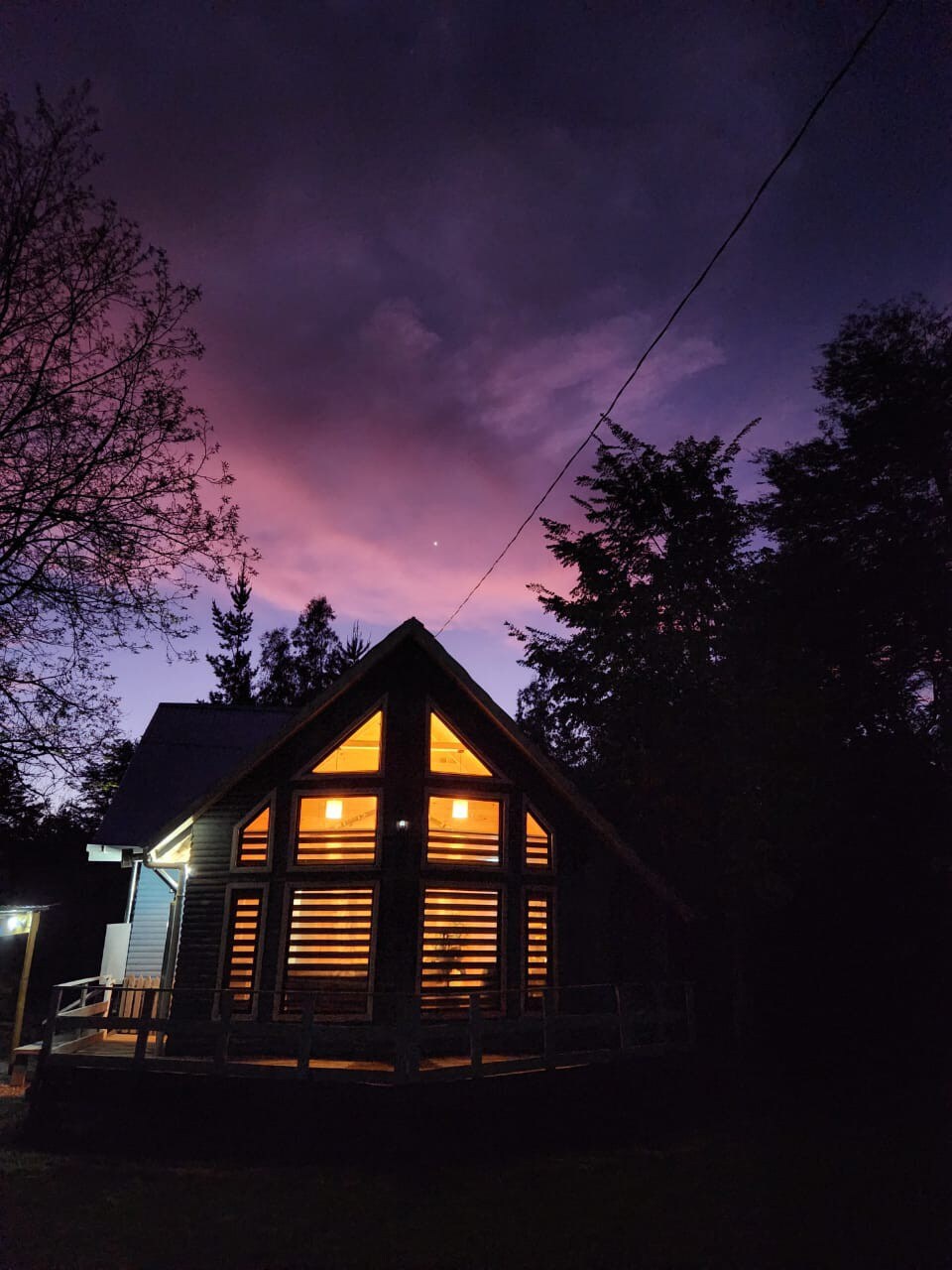 The exterior of the Cabaña Lodge Stone is showcased against a twilight sky, with warm light emanating from the windows. The structure features an A-frame design and a spacious deck. Surrounding trees are silhouetted against the vibrant colors of sunset, creating a serene atmosphere.