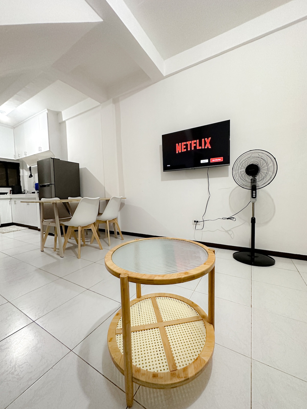 A living area features a mounted television displaying the Netflix interface. A round, two-tiered table stands on a tiled floor, accompanied by a dining table with four chairs in the background. A stand fan provides air circulation in the space.