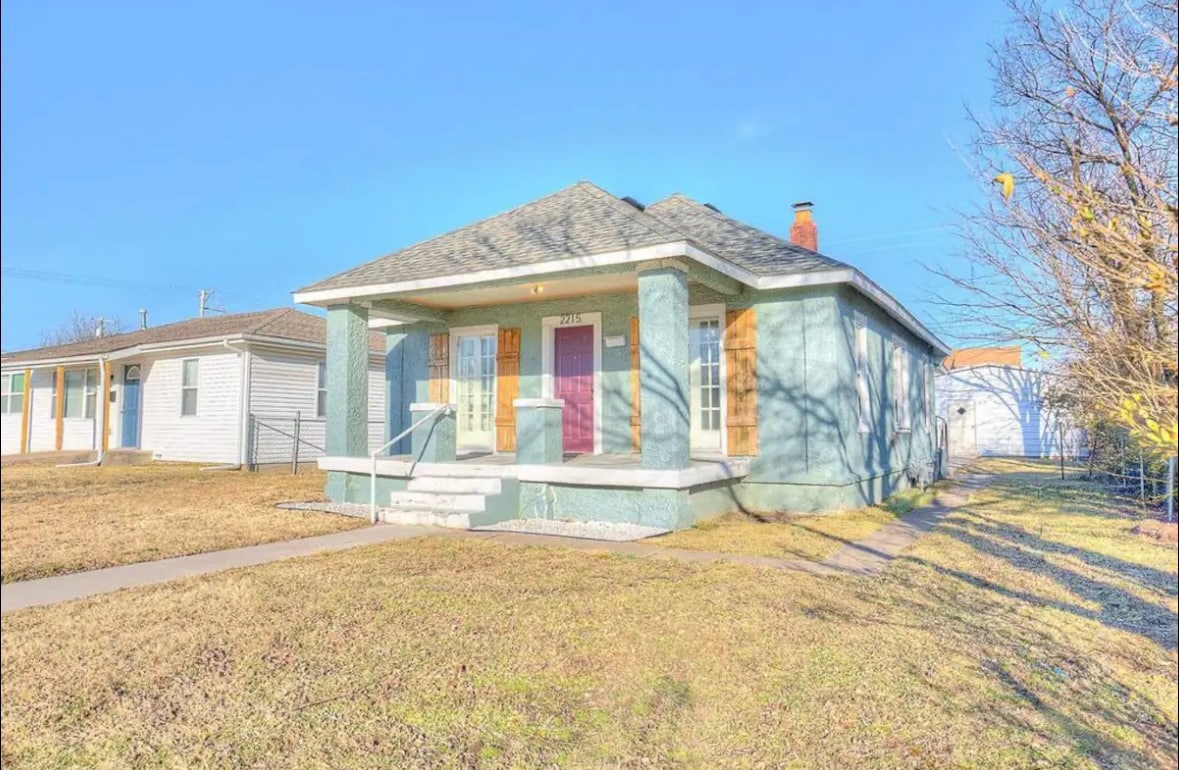 A charming single-story home is presented with a light blue exterior and a gabled roof. The front porch features steps leading to a central door, flanked by large windows. A well-maintained lawn is visible, bordered by a sidewalk and nearby houses.