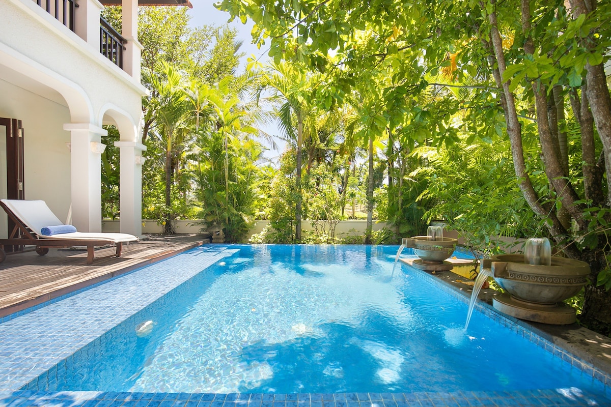 A serene pool area is surrounded by lush greenery, featuring calming water fountains on either side. A sunbed lies on the deck adjacent to the spacious pool, inviting relaxation under the dappled sunlight filtered through vibrant tropical plants.