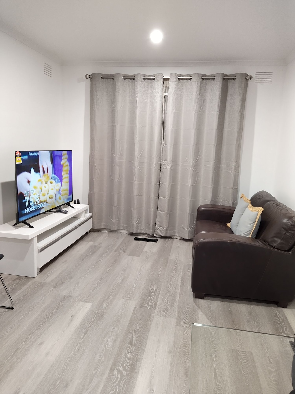 A living area features a dark leather sofa complemented by a decorative pillow. A flat-screen TV sits on a white console, displaying a vibrant image. Light-colored flooring and sheer curtains add a contemporary touch to the space.