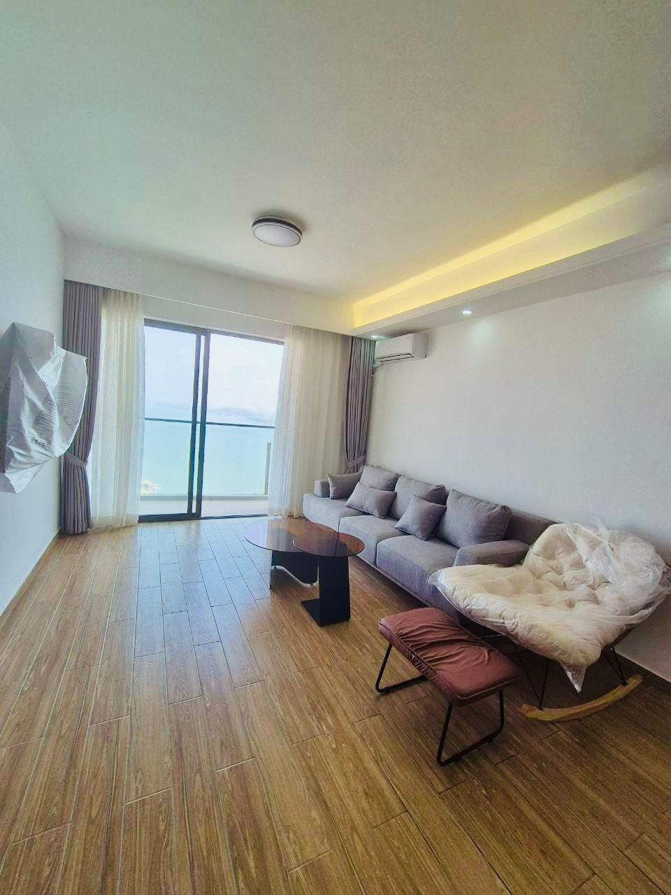 A modern living area features a comfortable gray sofa and a light-colored armchair. A round table provides a central focus, while large windows offer expansive views of the waterfront. Natural light brightens the space, complemented by soft curtain drapes.