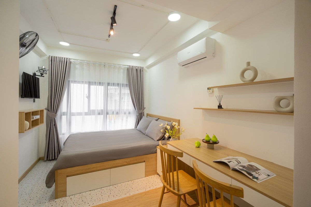 A modern sleeping area is equipped with a spacious bed dressed in gray linens. A small wooden table and two chairs offer a workspace. Large windows dressed with sheer curtains allow natural light to brighten the room, while a wall-mounted TV and decorative items add visual interest.