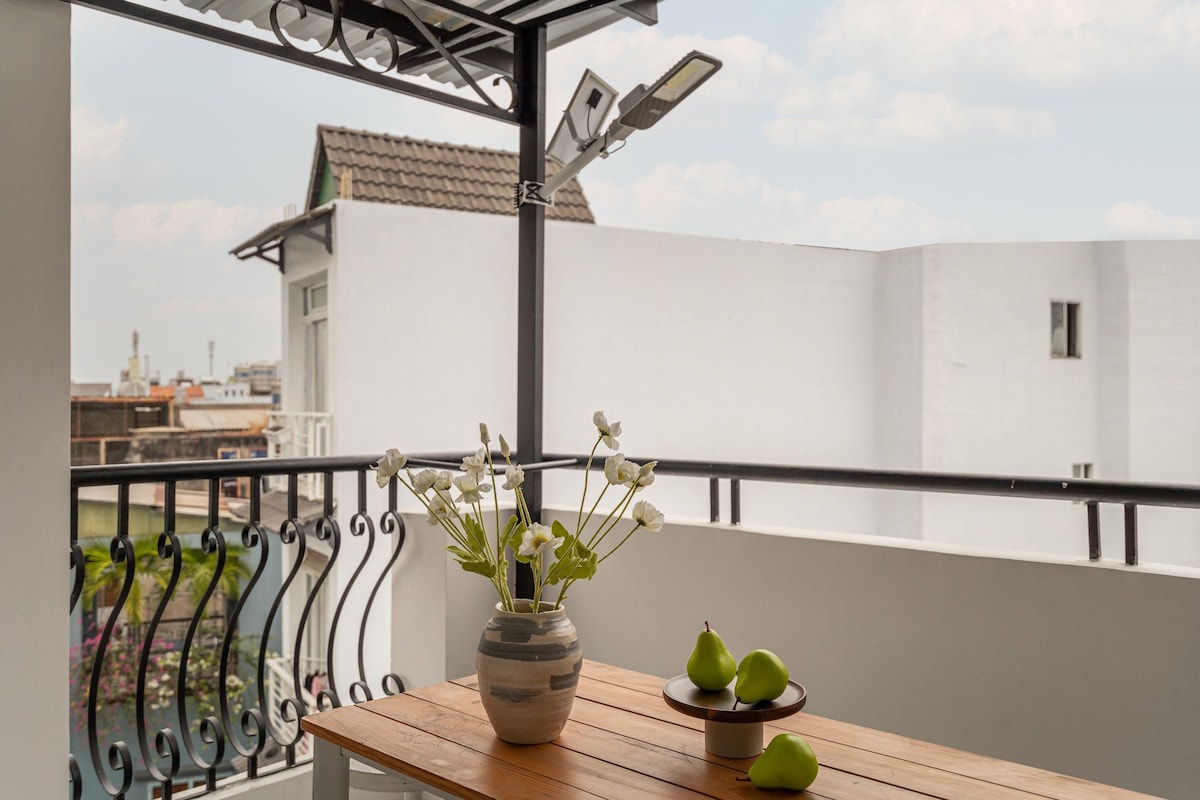 A balcony area features a wooden table set for outdoor relaxation, adorned with a vase of white flowers and a bowl of green fruit. The railing provides a view towards the skyline, complemented by a bright, open atmosphere.
