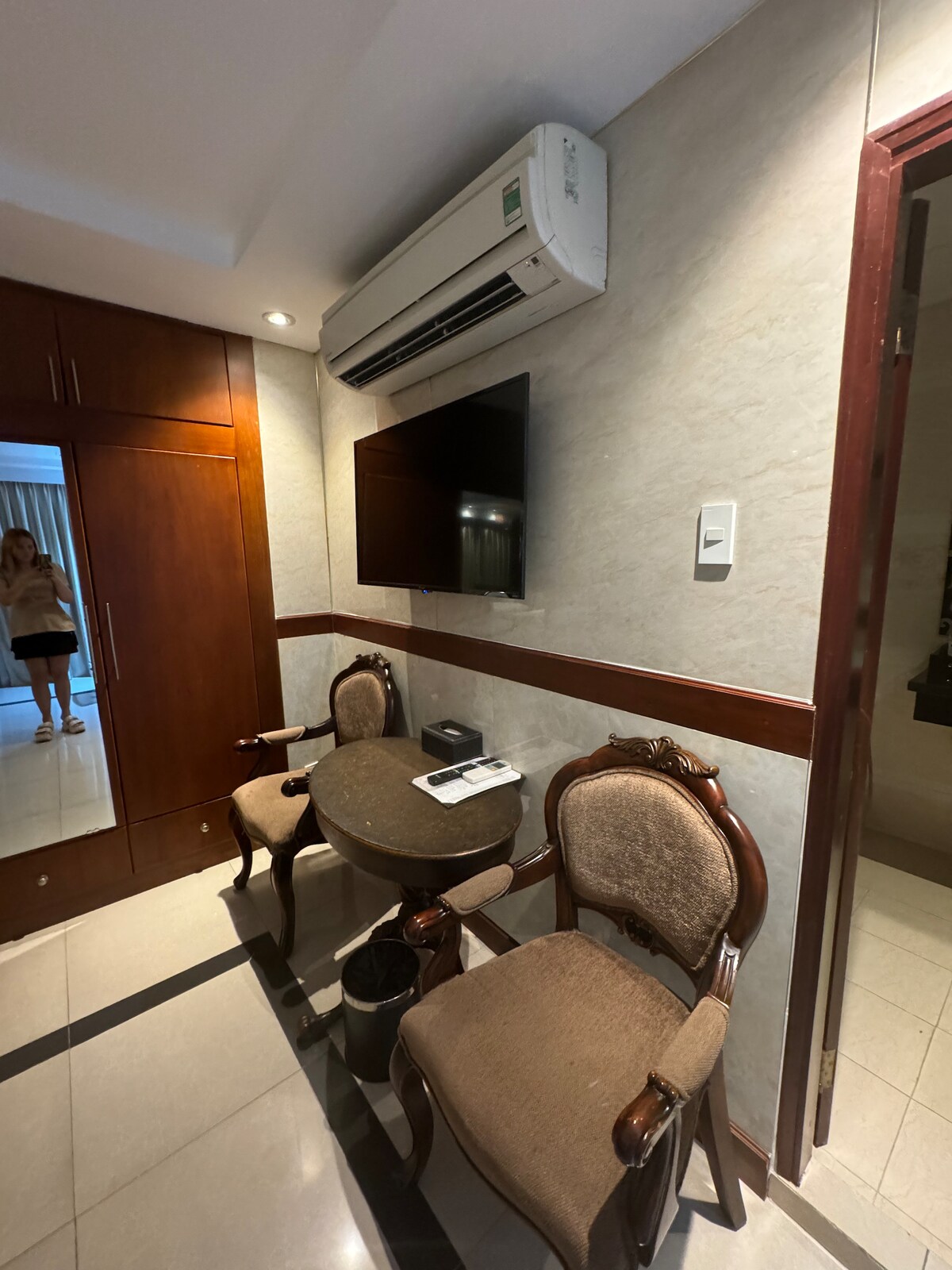 A small sitting area features a round table accompanied by two elegant chairs, set against a textured wall. A wall-mounted television is situated above the table, while a mirrored wardrobe stands nearby, reflecting some natural light from an adjacent window.