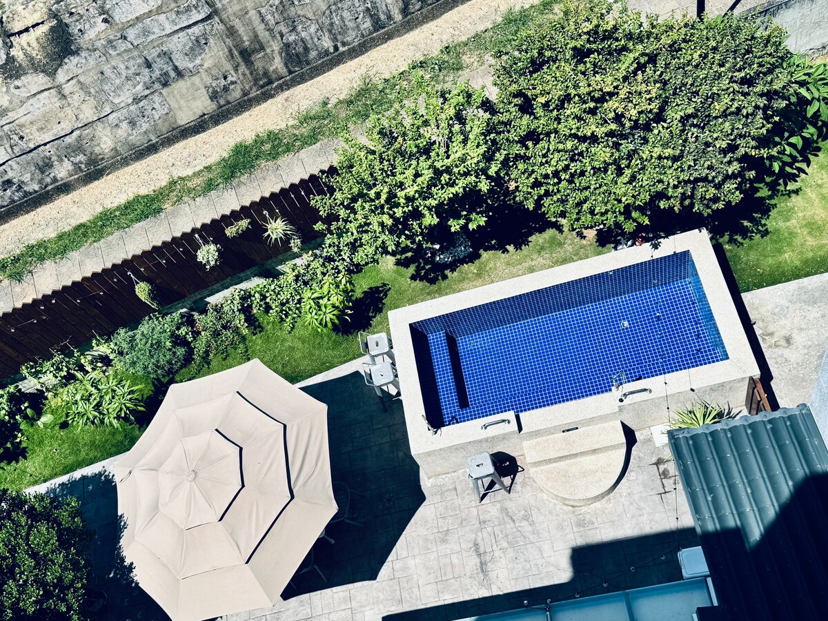 An elevated view captures a private outdoor area featuring a rectangular swimming pool surrounded by lush greenery. A shaded patio area with an umbrella and seating is visible nearby, along with carefully landscaped plants enhancing the tranquil setting.