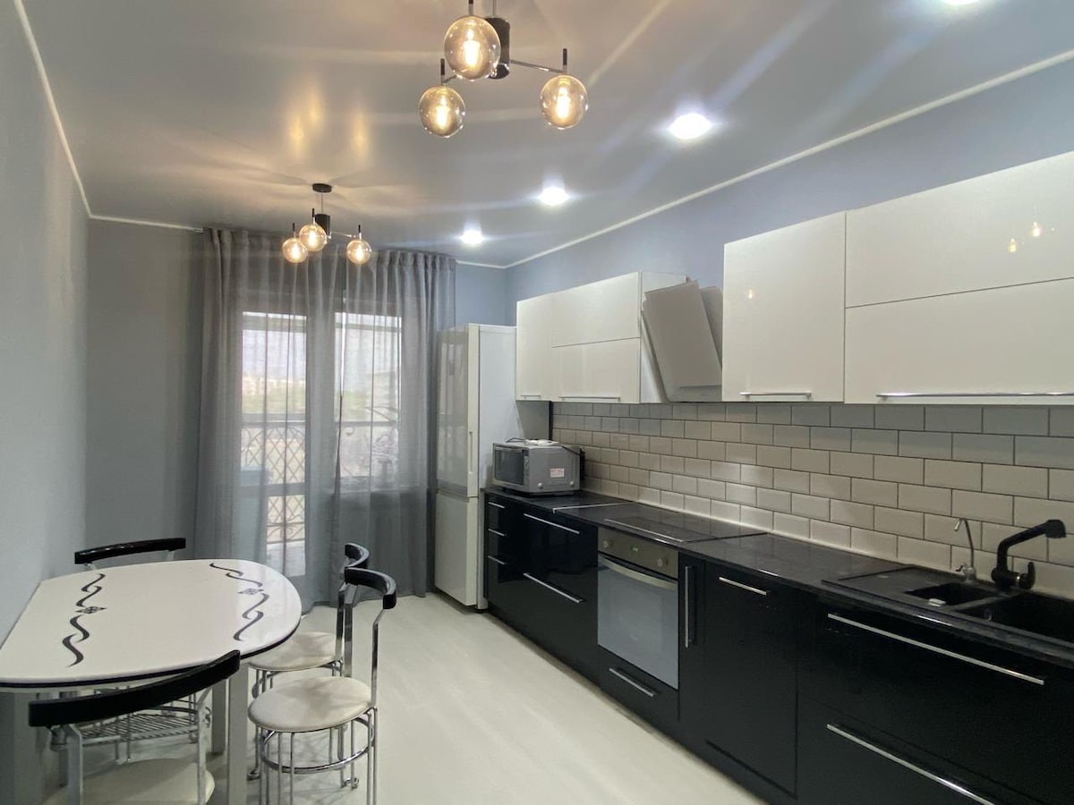 A modern kitchen is featured with sleek black cabinets and white countertops. An inviting dining table with two chairs is positioned near a window, allowing natural light to stream in through sheer curtains. Stainless steel appliances add a functional touch to the space.