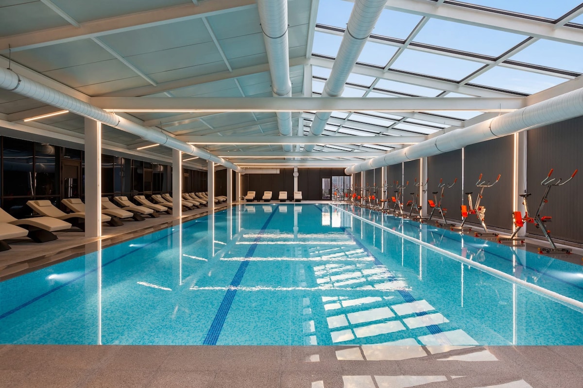 A spacious indoor pool area is illuminated by natural light from overhead skylights. Comfortable lounge chairs line one side, while exercise bikes are positioned along the opposite side. The clear blue water reflects the bright surroundings, creating a serene atmosphere for relaxation and fitness.