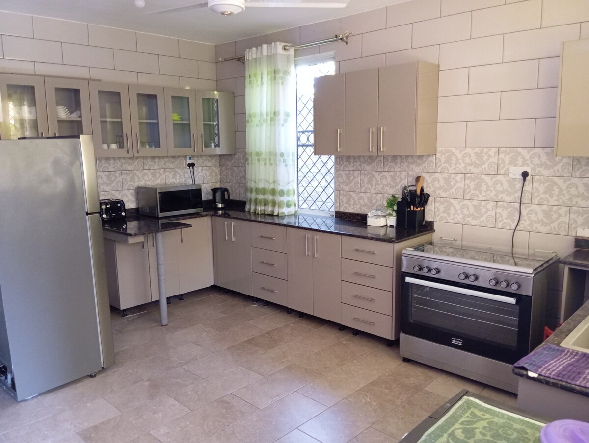 A modern kitchen features sleek cabinetry in neutral tones, paired with a dark granite countertop. Essential appliances, including a stainless steel refrigerator, microwave, and oven, are visible. Natural light enters through a window, complemented by light sheer curtains.
