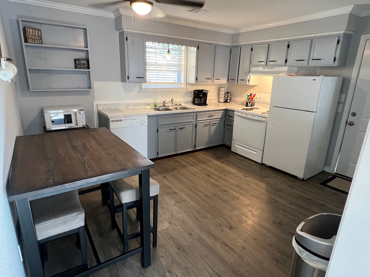 A functional kitchen area is presented with modern grey cabinetry and white countertops. Equipped with essential appliances, including a refrigerator, stove, and microwave, it includes a small dining table with four chairs. Natural light enters through a window, enhancing the workspace.