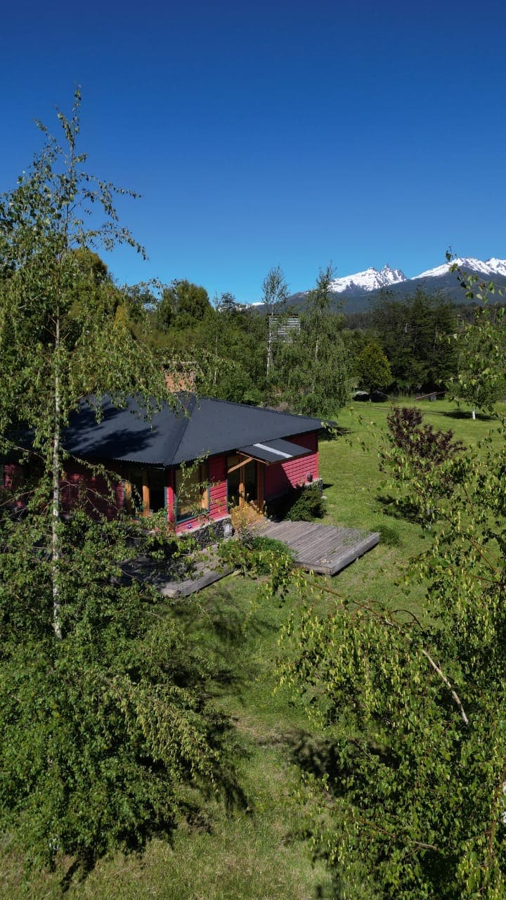 The exterior of the cabin is framed by lush greenery and trees, with a wooden deck extending from the entrance. Snow-capped mountains are visible in the distance against a clear blue sky.