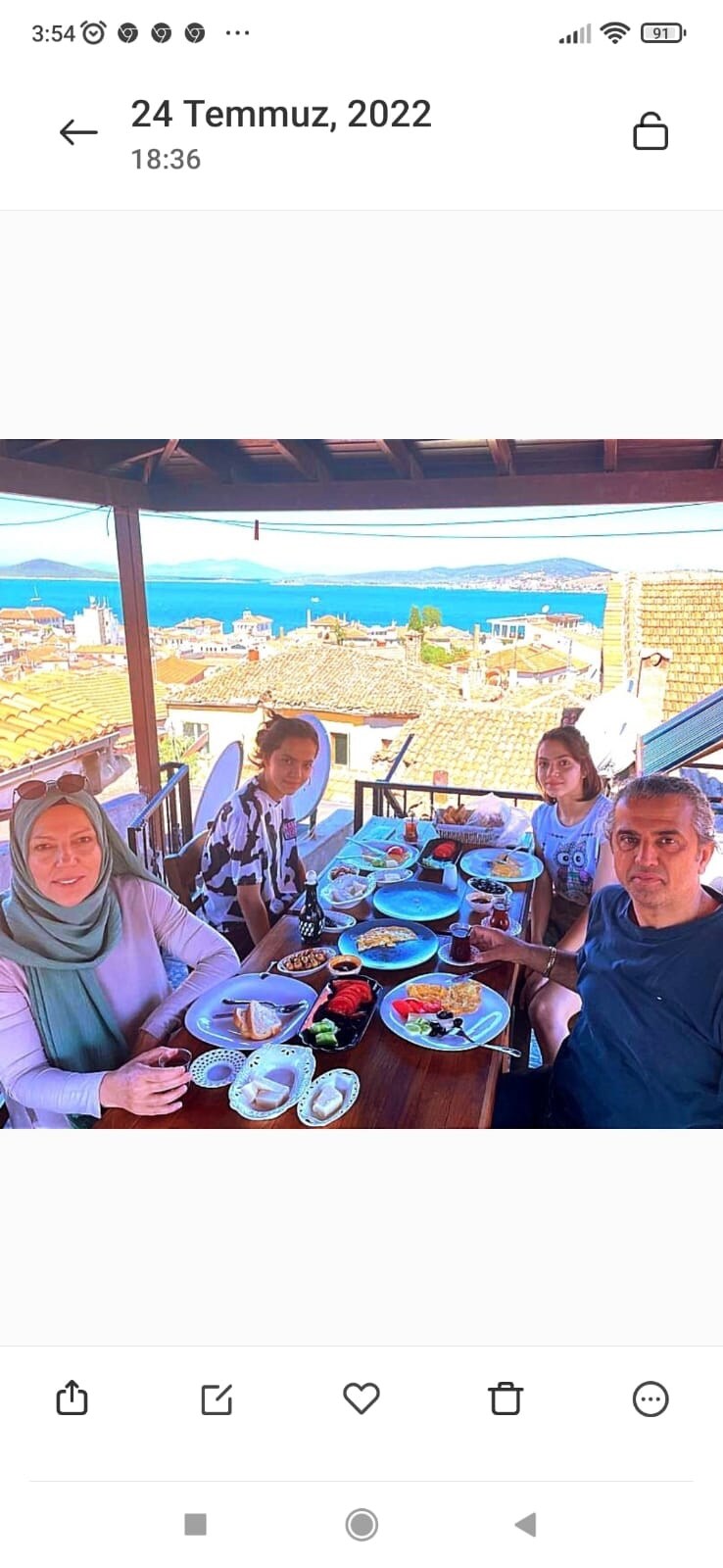 A wooden table is set with various plates of food, featuring fresh fruits and pastries. Several glasses are placed nearby. In the background, a scenic view of the town and sea can be seen under a clear sky. The setting suggests a relaxed dining experience on a terrace.