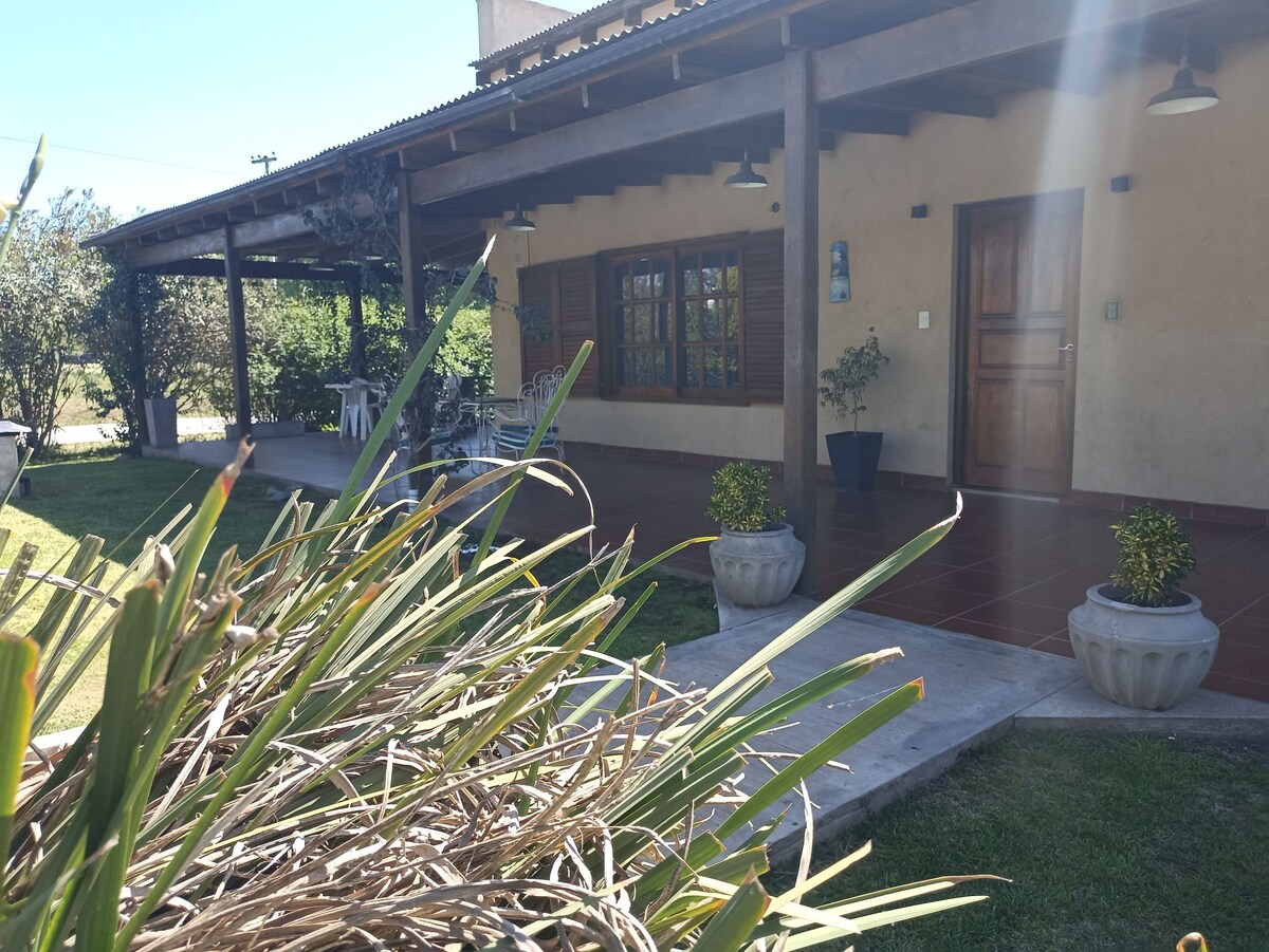 A welcoming facade of a home is displayed, featuring a covered porch with wooden beams. The entrance door is flanked by a small potted plant and windows with wooden shutters. A grassy area surrounds the entrance, adding to the outdoorsy ambiance.